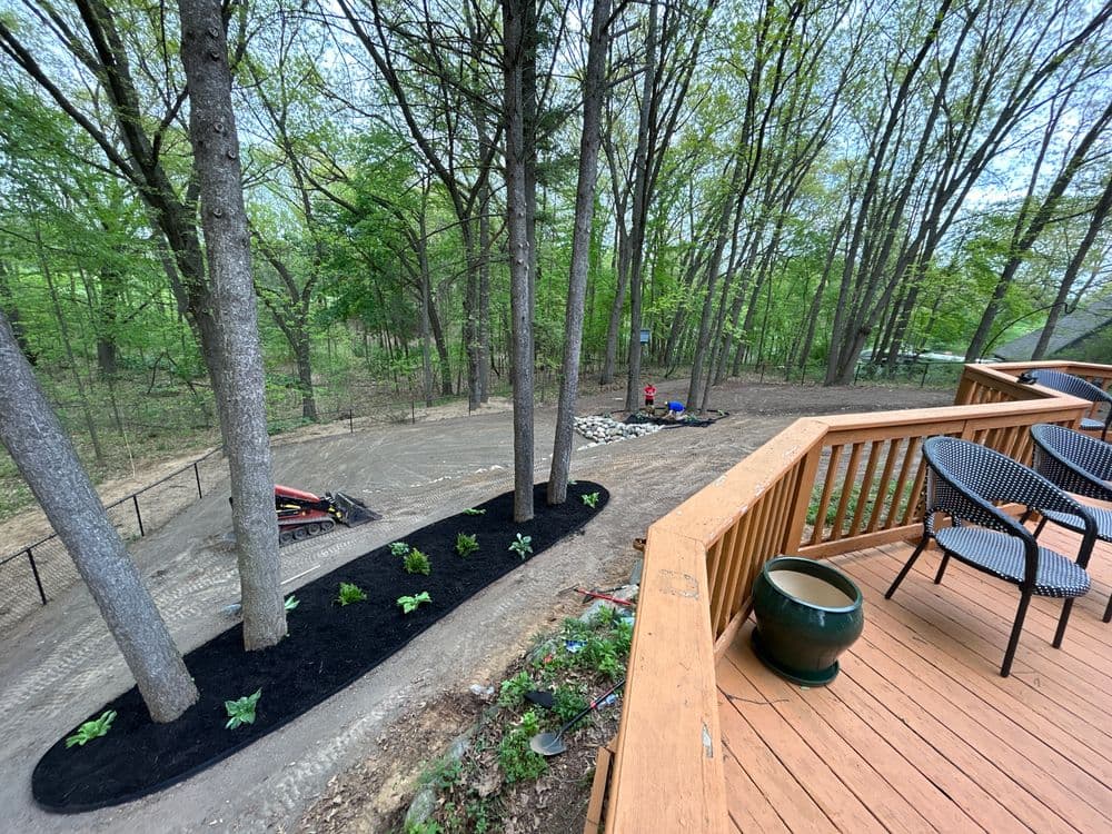View from a deck overlooking a landscaped area with trees, plants, and machinery in a wooded setting.