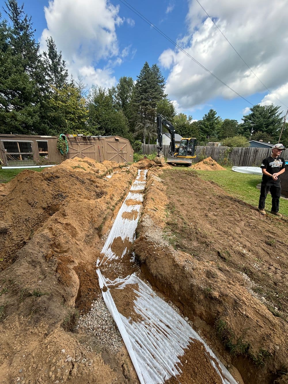 Excavation site with a trench and drainage fabric, machinery in background, trees nearby.